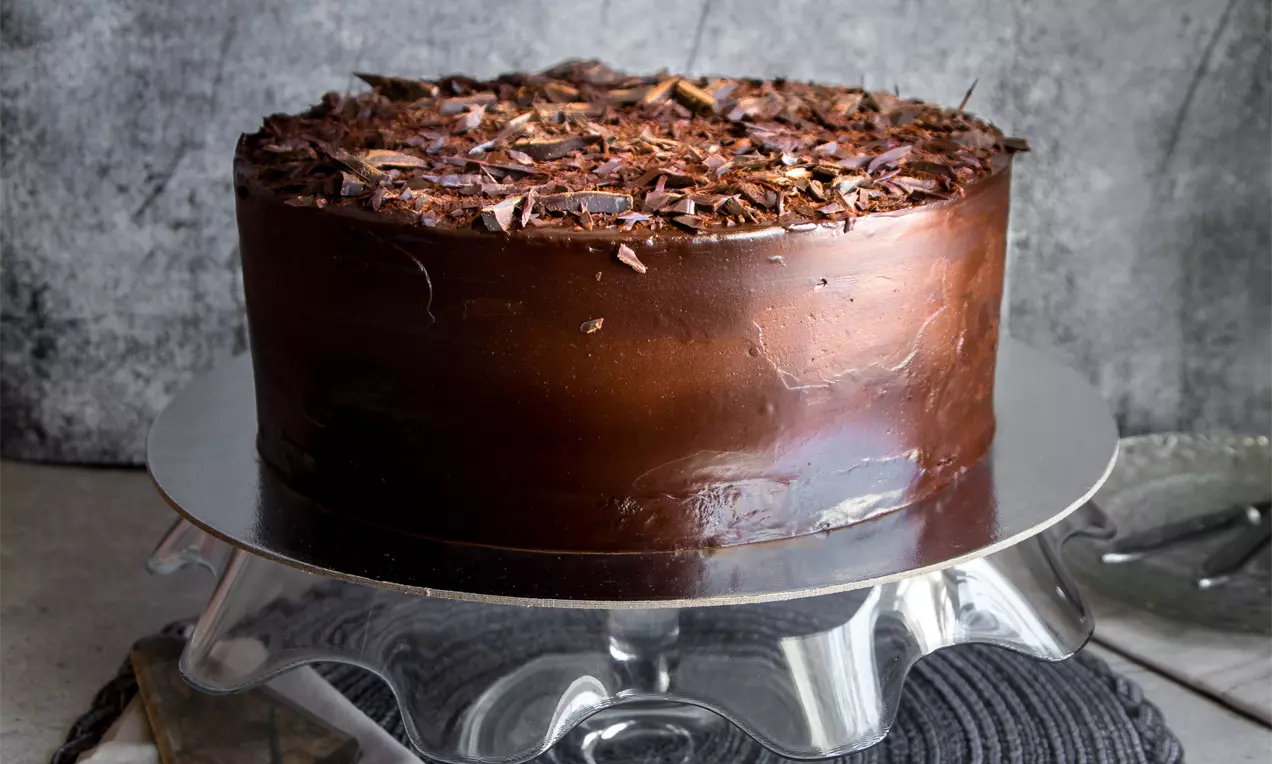 Кръгъл шоколадов сладкиш с плътен тъмен ганаш, украсен с едри шоколадови стърготини върху стъклена стойка на текстурирана основа