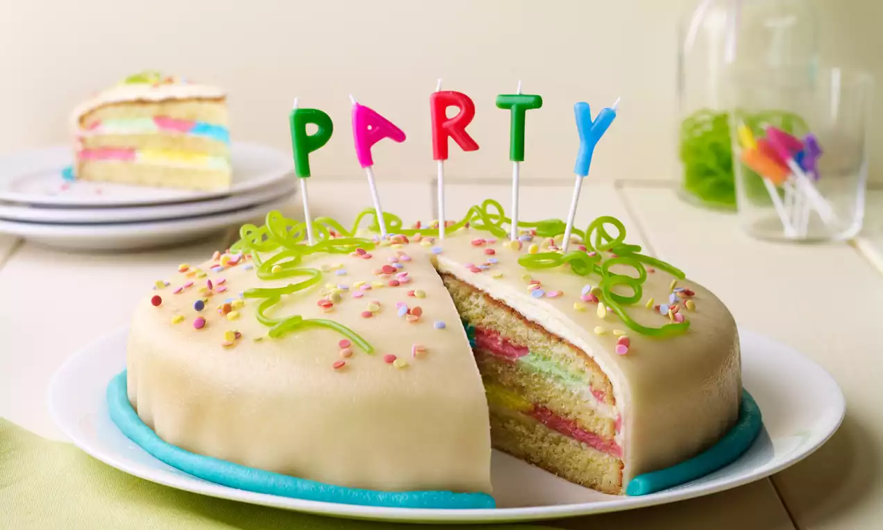 Vanillefarbene Torte mit bunter Füllung und Dekor aus grünen Zuckerspiralen, bunten Streuseln und farbigen PARTY-Kerzen auf weißer Platte