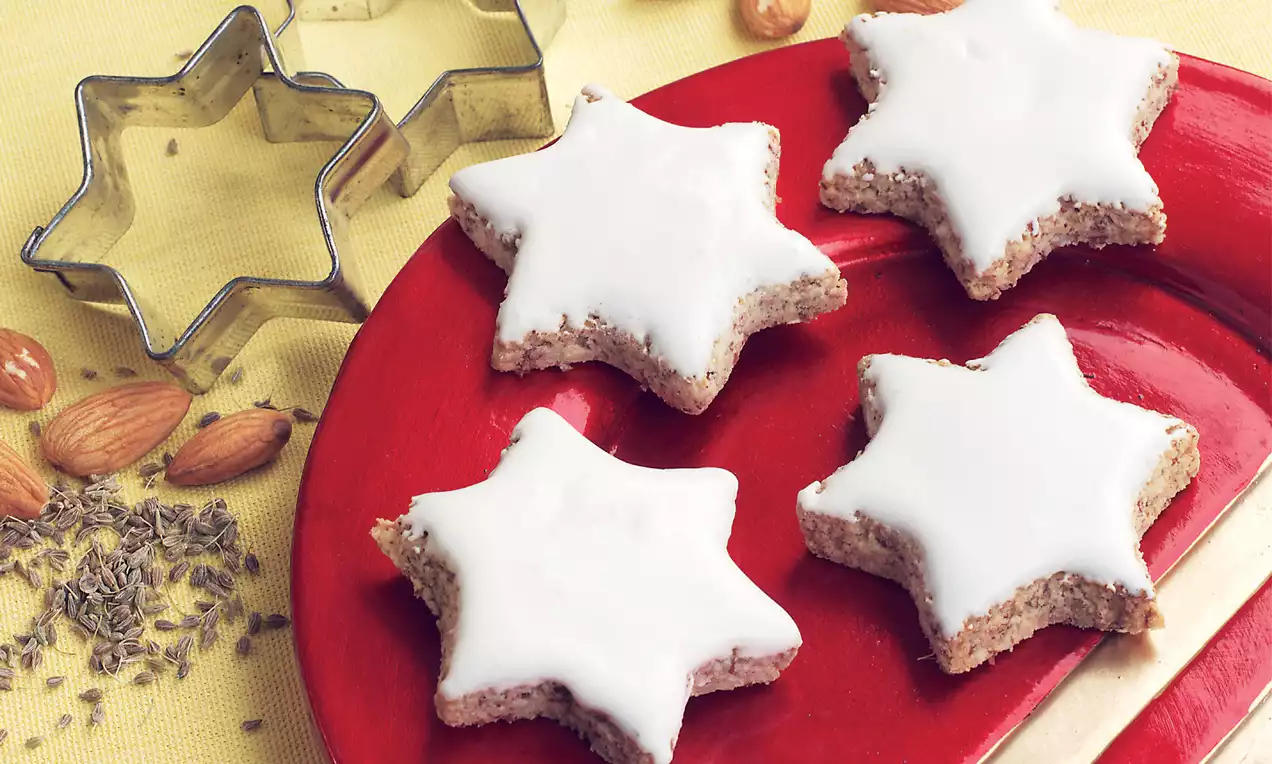 Weiße Anissterne mit zarter Glasur und krümeligem Rand auf rotem Teller, daneben Anis, Mandeln und sternförmige Ausstecher