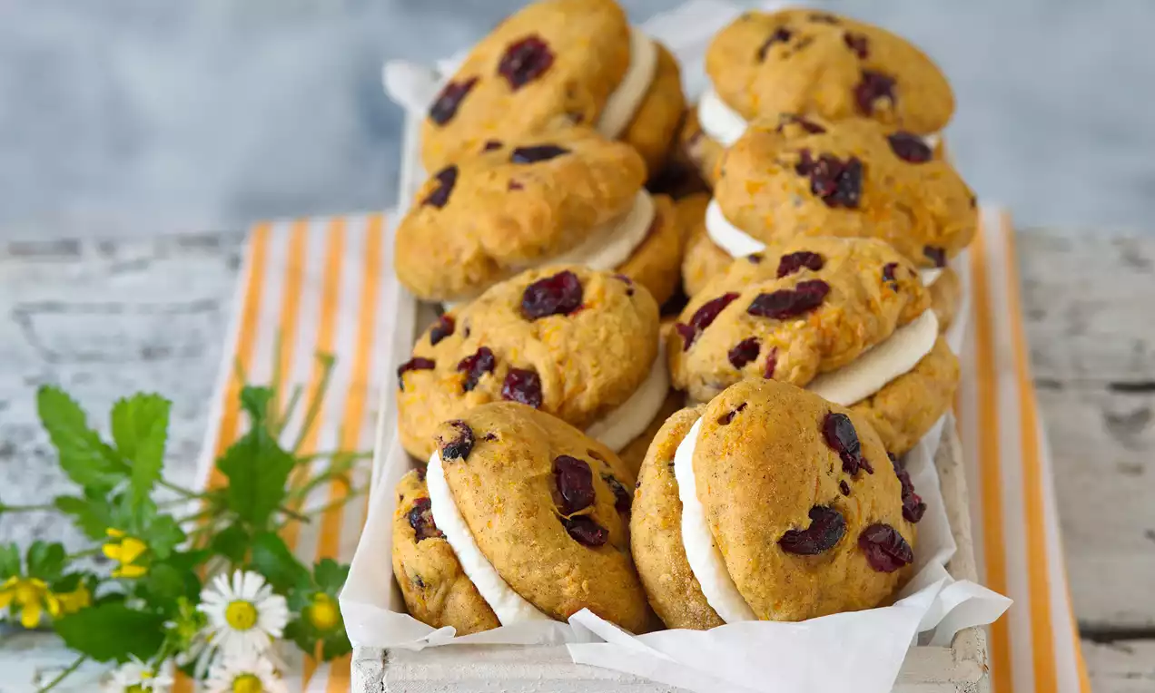 Mrkvové cookies světle oranžové barvy s kousky sušených brusinek, spojené vrstvou bílého krému a naaranžované v dřevěné bedýnce na pruhovaném ubrousku