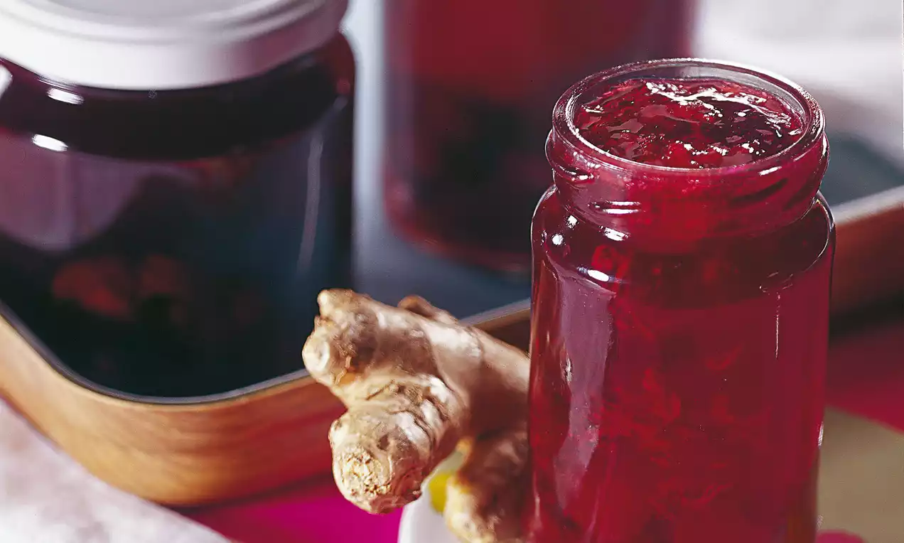 Ein Glas mit glänzend roter Sauerkirschkonfitüre, grobe Fruchtstücke sichtbar, daneben frischer Ingwer auf heller Unterlage drapiert