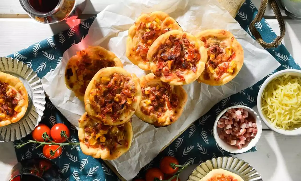 Goldbraun gebackene Pizza-Muffins mit knusprigem Rand und Käse, angerichtet auf Backpapier, umgeben von Tomaten, Reibekäse und Speckwürfeln