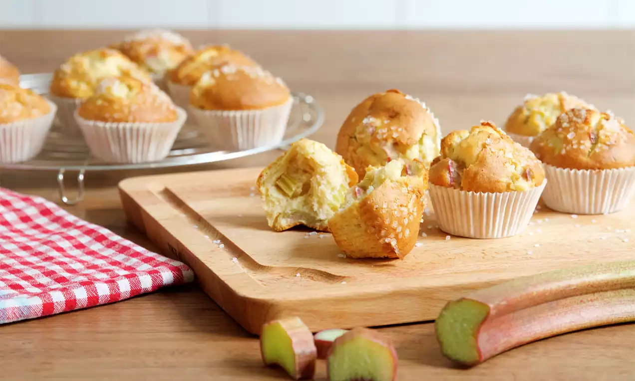 Goudbruine rabarbermuffins in witte papiertjes op een houten plank, met rabarberstengels en een rood-wit geblokt doek op de achtergrond