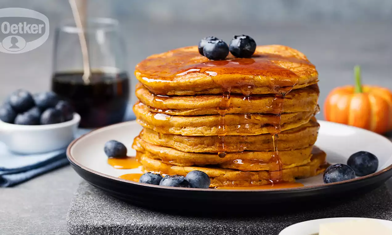 Tour de pancakes dorés à la citrouille nappés de sirop ambré, parsemés de myrtilles, sur une assiette blanche avec une mini citrouille