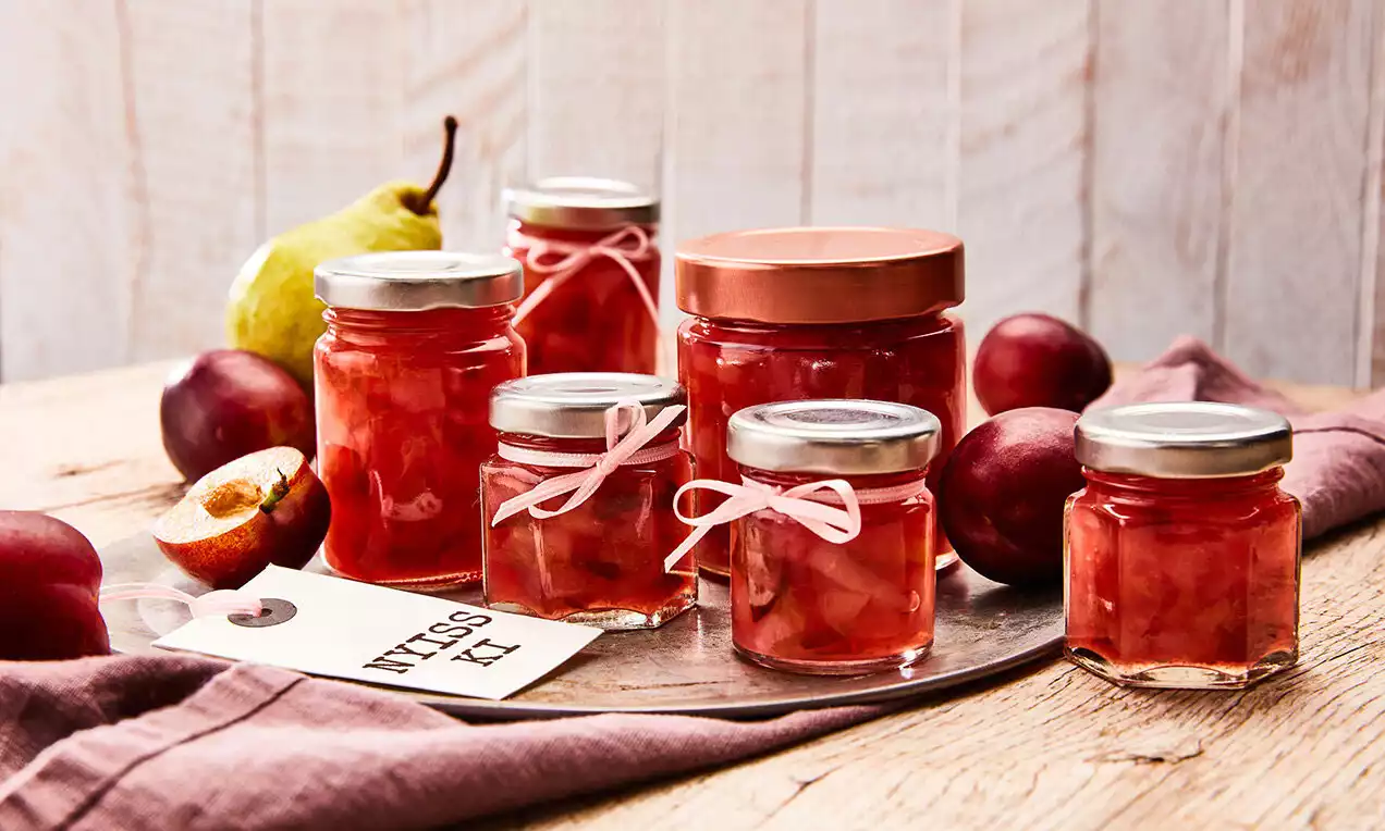Üvegekben mély bordó dzsem, áttetsző rétegben a gyümölcshús darabjaival, körülöttük szilvák és körte natúr fa tálcán, puha textillel