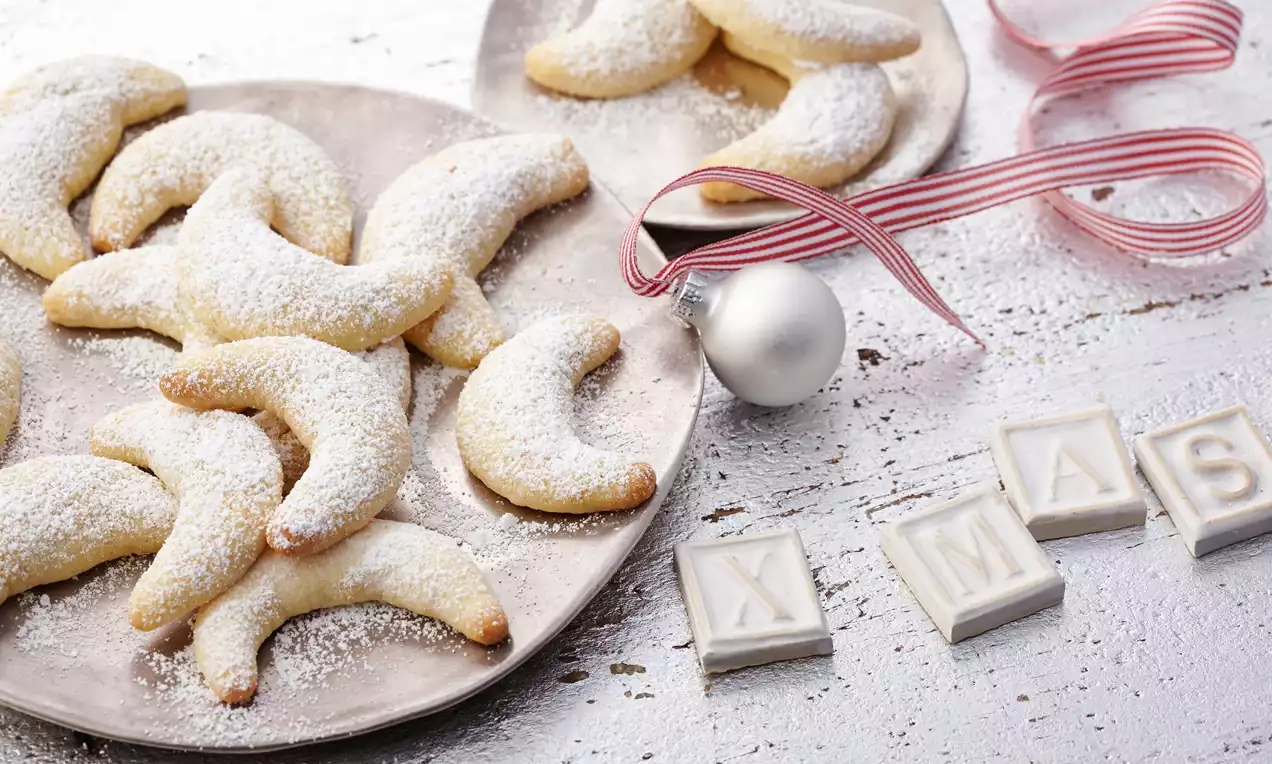 Vanillekipferl en forme de croissants courbés saupoudrés de sucre glace sur une assiette argentée avec une boule blanche et ruban rayé