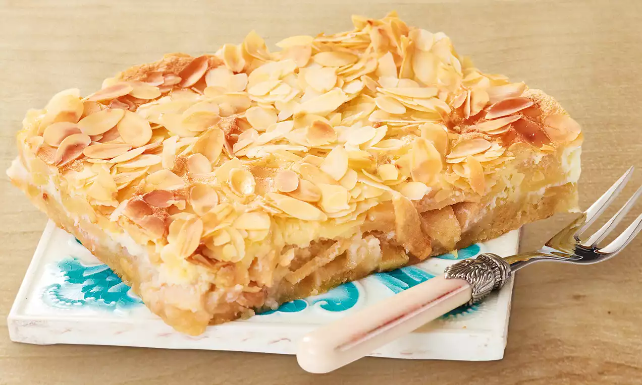 Carré de gâteau doré aux pommes et amandes effilées sur une assiette décorée aux motifs bleus, accompagné d'une fourchette à manche clair