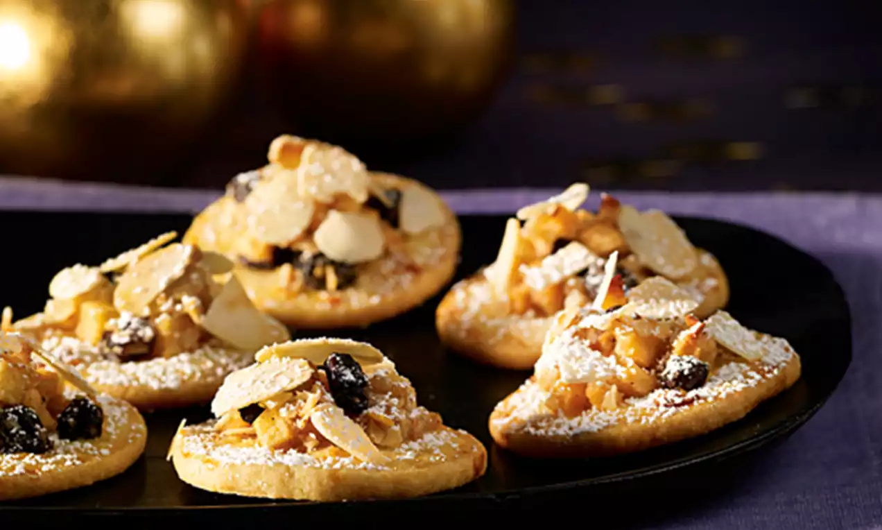 Bratapfel-Plätzchen mit goldgelbem Teig, bestrichen mit Apfelstückchen, Rosinen und Mandelblättchen, leicht mit Puderzucker bestäubt auf schwarzem Teller