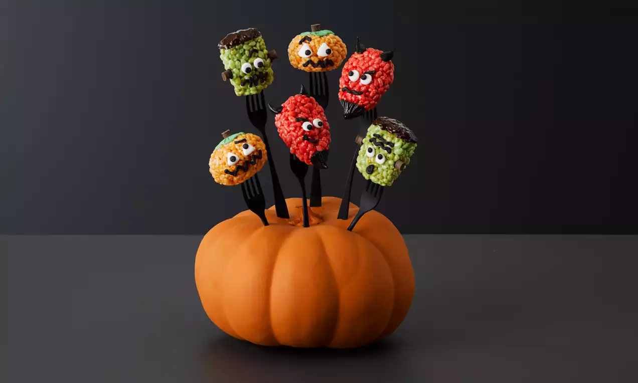 Vibrant orange pumpkin holding black forks topped with round marshmallow crispies in green, orange, and red with playful monster faces