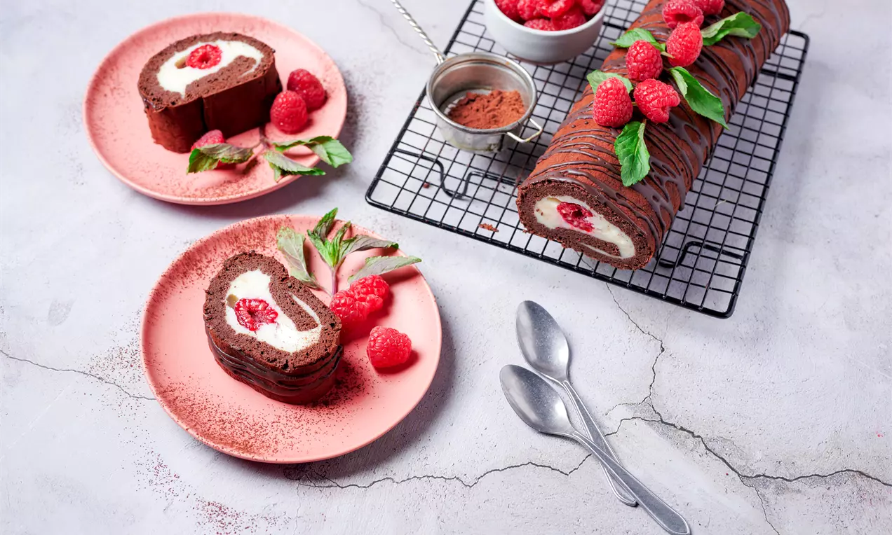 Шоколадово руло с кремообразен бял пълнеж и малиново сърце, обвито в тънки шоколадови ленти, сервирано с пресни малини и листа върху решетка и розови чинии