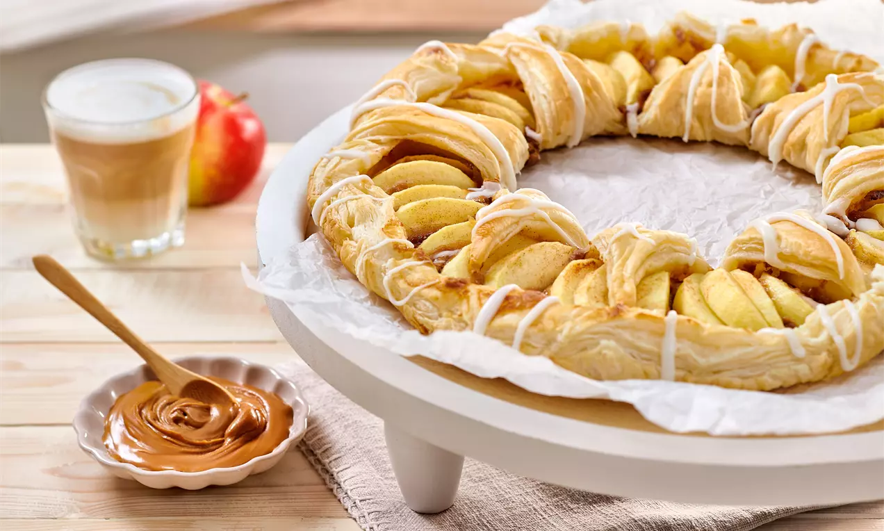 Runde Apfel-Tarte aus goldenem Blätterteig mit frischen Apfelscheiben und weißem Zuckerguss auf einem weißen Teller mit Lotuscreme-Schale