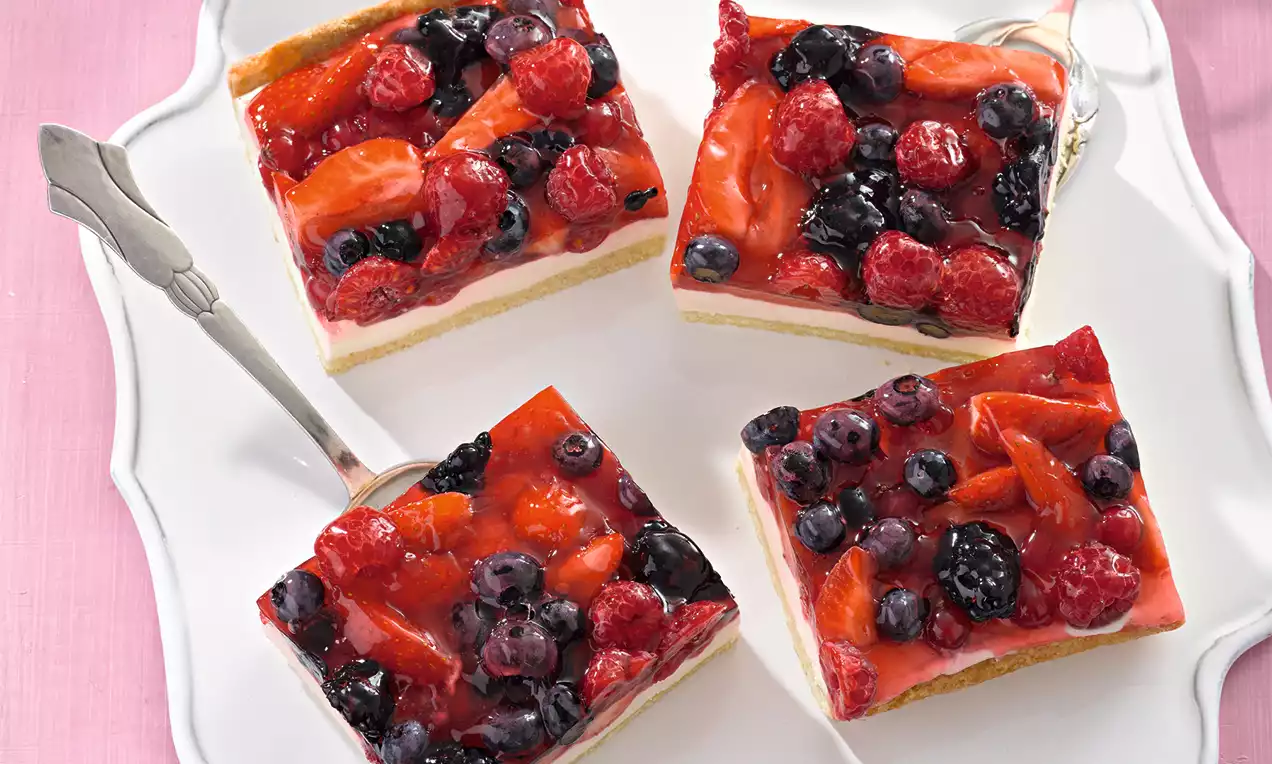 Čtvercové řezy se světlým piškotem, bílou zakysanou smetanou, lesklou želatinou a šťavnatým ovocem na bílém talíři s dekorativním okrajem