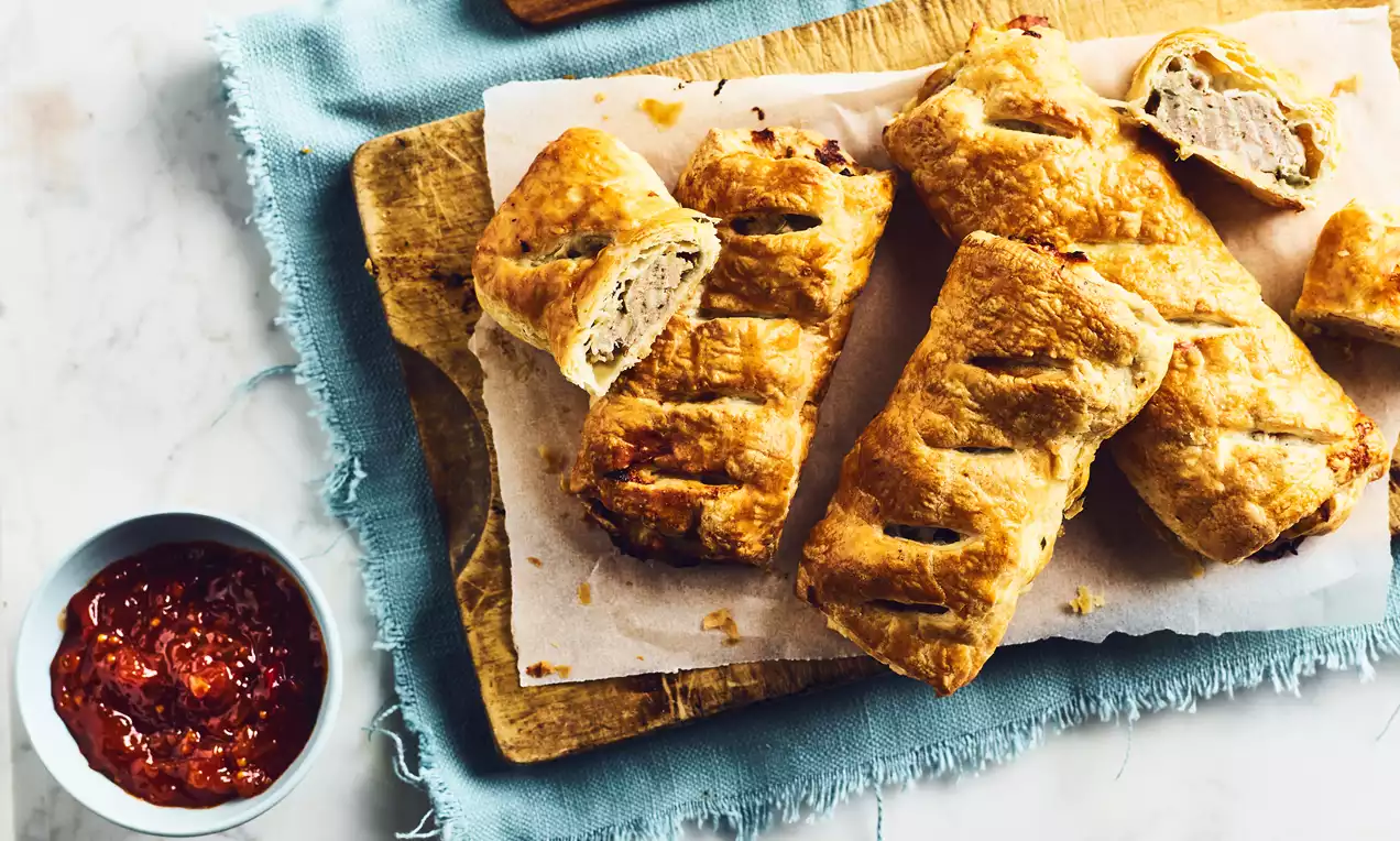 Golden, flaky sausage rolls with diagonal slits, arranged on parchment atop a wooden board, served with a bowl of red chutney