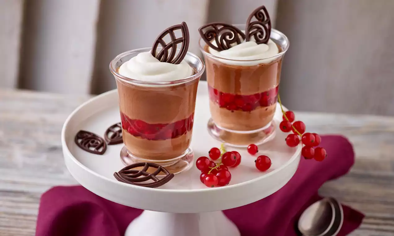 Zwei Gläser mit Schoko-Mousse, roter Fruchteinlage und Sahnehäubchen, dekoriert mit filigraner Schokogarnitur, auf weißer Tortenplatte