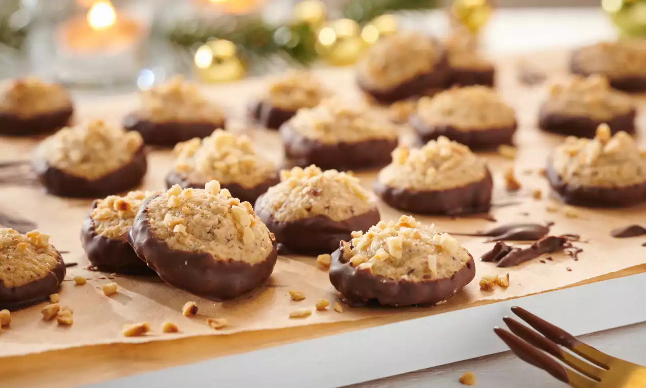 Marzipan-Makronen mit goldbraunem Haselnusskern und dunkler Schokoglasur auf Backpapier, umgeben von dekorativen Weihnachtslichtern