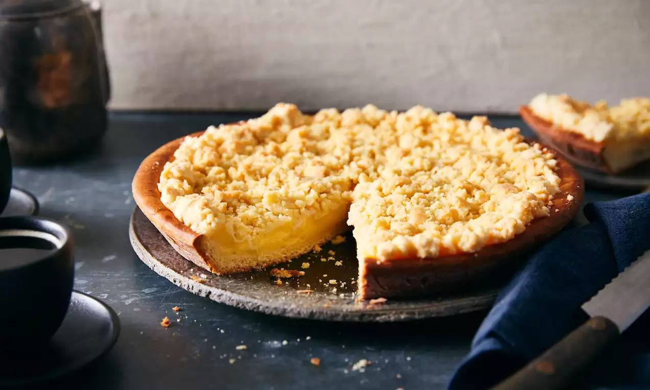 Tarte ronde dorée avec une base moelleuse, garnie de crème pâtissière jaune et d’un crumble croustillant, sur une assiette en pierre