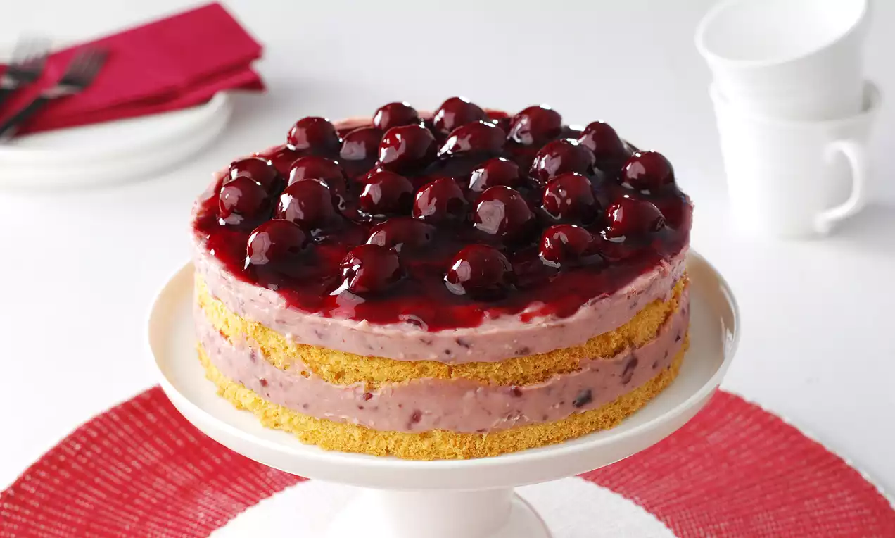 Altın sarısı pandispanya katları arasında vişneli pastacı kreması, üstte parlak vişne sosu ve kibar bir sunumla beyaz ayaklı tabakta