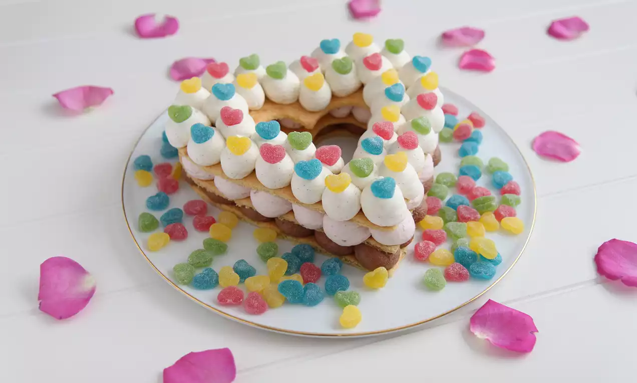 Hjerteformet kake med lyse kremtopper og fargerike geléhjerter på hvitt fat med gullkant, omgitt av rosa roseblader