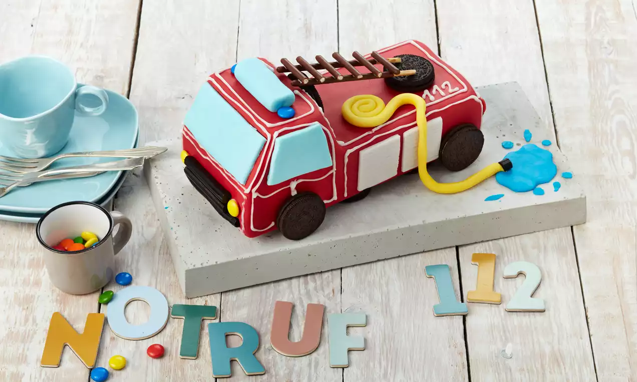 Feuerwehrauto-Kuchen mit glänzend rotem Zuckerguss, schwarzen Fondant-Reifen, silberner Leiter, gelben Scheinwerfern und bunten Kerzen