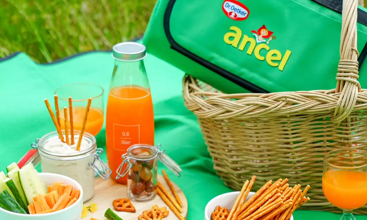 Bouteille et verre de jus orange brillant avec nuances carotte et citron, posés sur une nappe verte près de snacks et légumes croquants