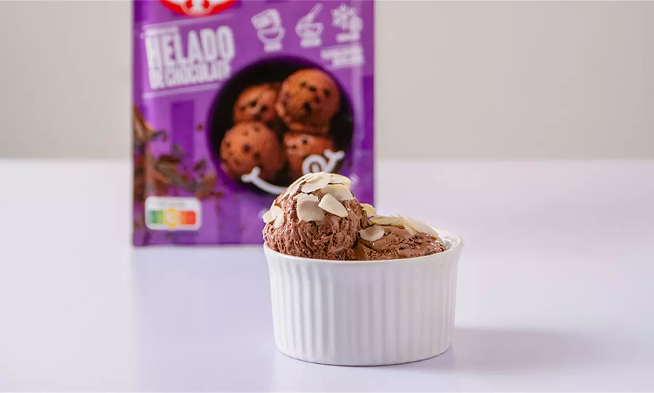 Helado de chocolate marrón en bolas suaves dentro de un recipiente blanco, decorado con láminas de almendra sobre fondo morado