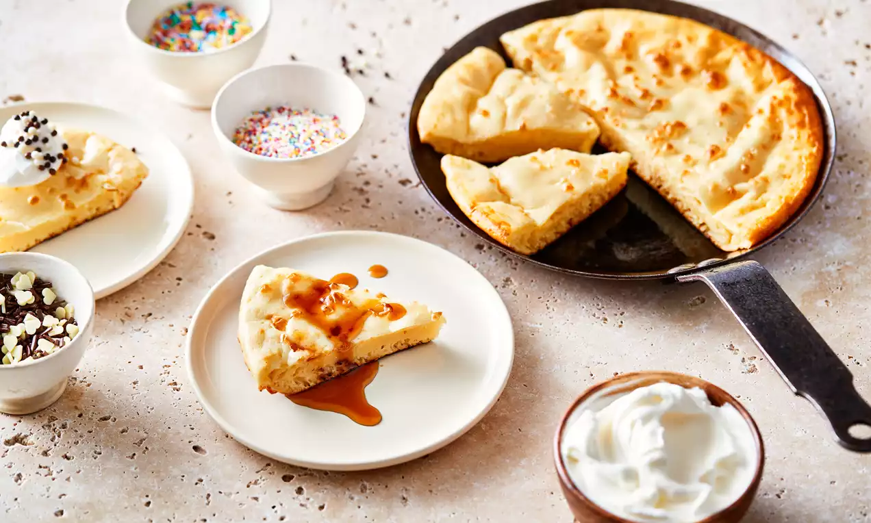 XL pannenkoek goudbruin en luchtig in een zwarte pan, met gesneden stukken op witte borden en karamelsaus, omgeven door schaaltjes decoratie