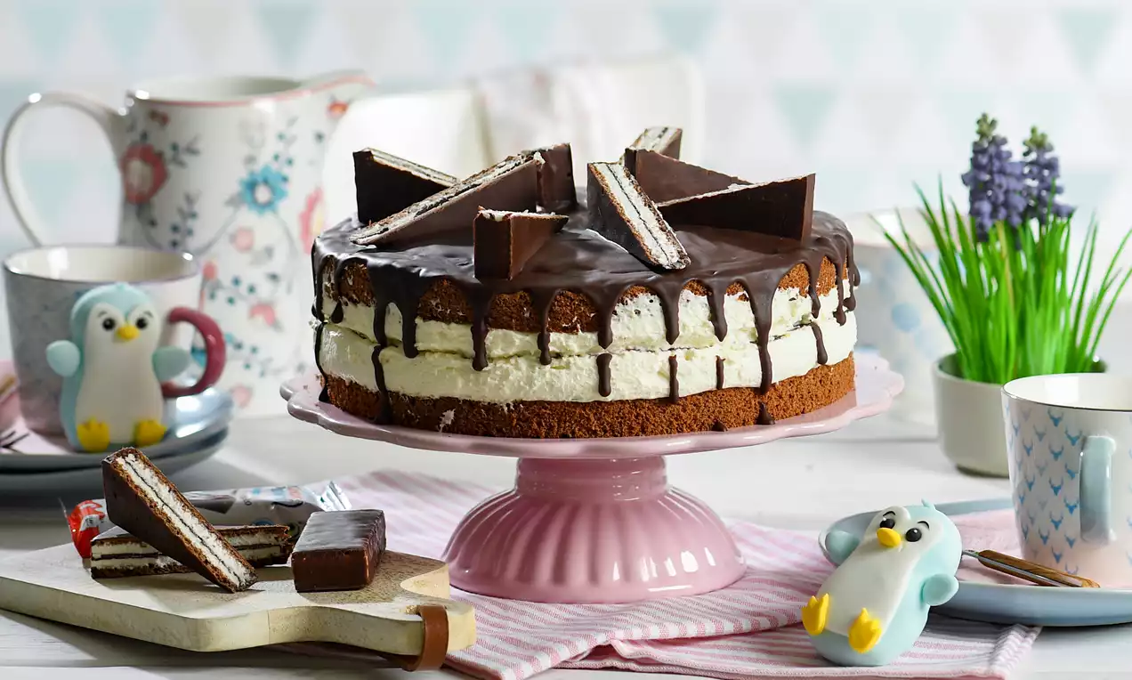 Schokoladentorte mit weißer Cremefüllung, dunklem Schokoguss und Kinder-Pingui-Stücken, auf rosa Tortenplatte, umgeben von Dekoration