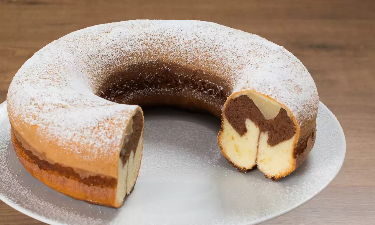 Ciambella bicolore soffice, con strati alternati di impasto bianco e al cacao, spolverata di zucchero a velo su un piatto bianco