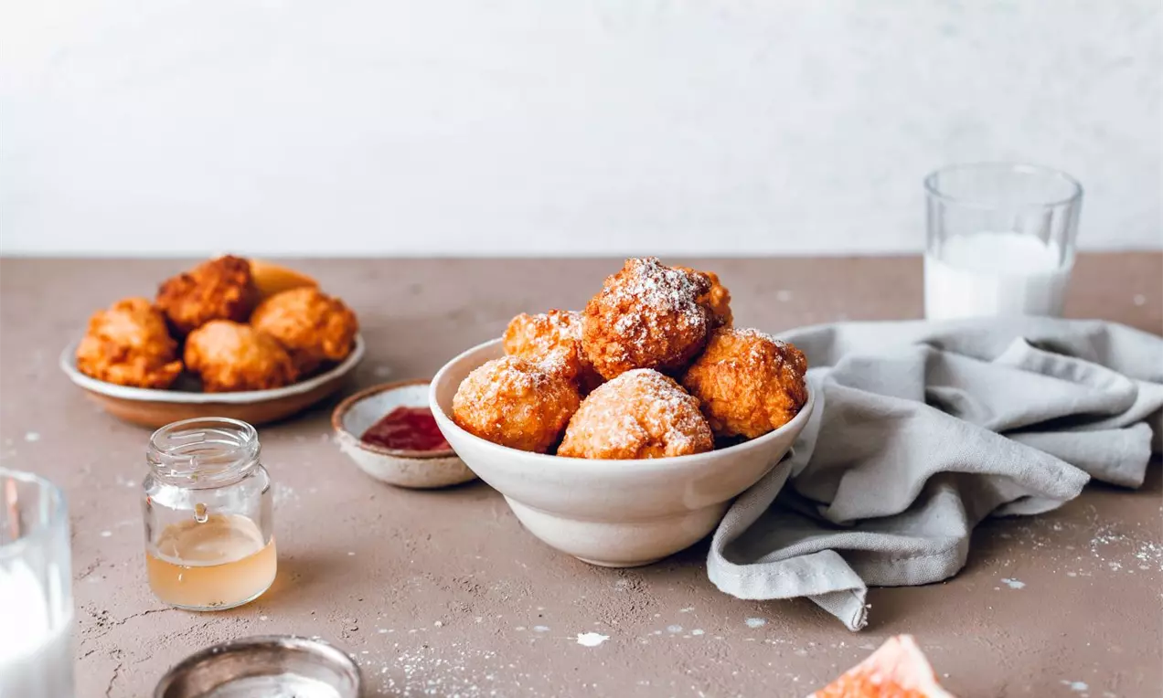 Zlatohnedé guľaté múčne guličky, jemne posypané práškovým cukrom, usporiadané v keramickej miske na hnedom stole vedľa šedej utierky