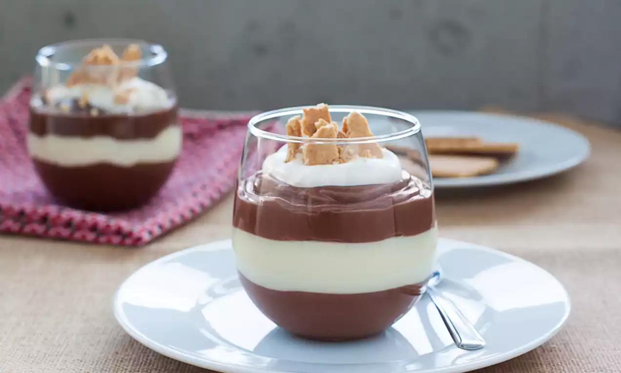 Pudding en verrine à deux couches chocolat et vanille surmonté de crème fouettée blanche et éclats de biscuit dorés sur assiette blanche