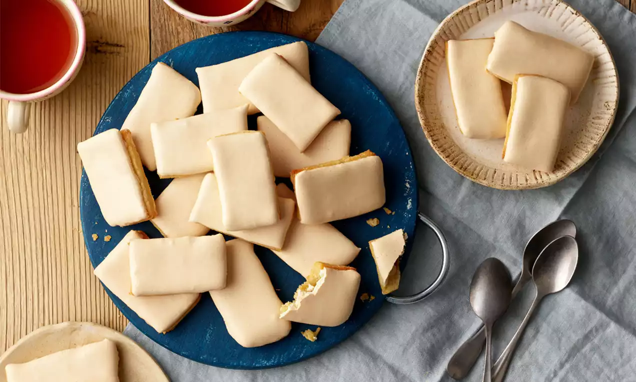 Butterstangerl mit zartem, hellbeigem Überzug, rechteckig und glatt, locker arrangiert auf blauem Teller neben Tee und Löffeln