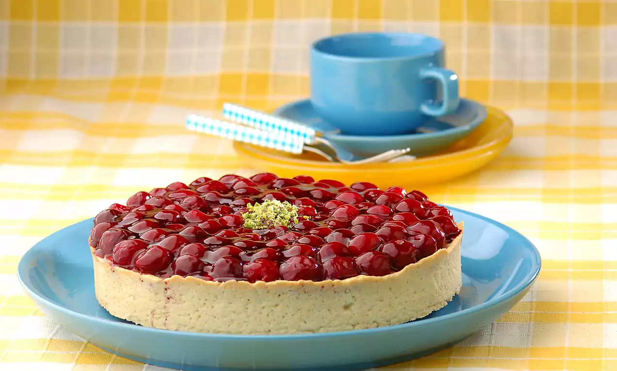Altın renkli gevrek tart tabanı üzerine kremalı dolgu, parlak kırmızı vişne taneleri ve ortasında ince kıyılmış yeşil fıstık ile süslenmiş
