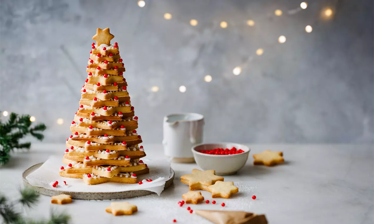 Picture - A cookie Christmas tree of stacked star-shaped biscuits in golden tones with white icing and red sprinkles, set on parchment paper