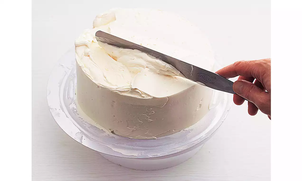 Runde Torte auf weißer Platte, glatt und deckend mit cremefarbener Buttercreme bestrichen, Hand streicht Oberfläche mit einer Palette