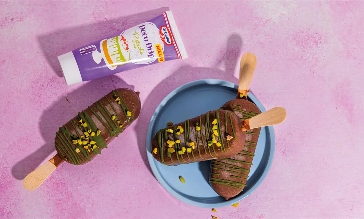 Donkerbruine chocolade-ijsjes op houten stokjes, besprenkeld met groene pistachenoten, op een blauw bord en roze ondergrond