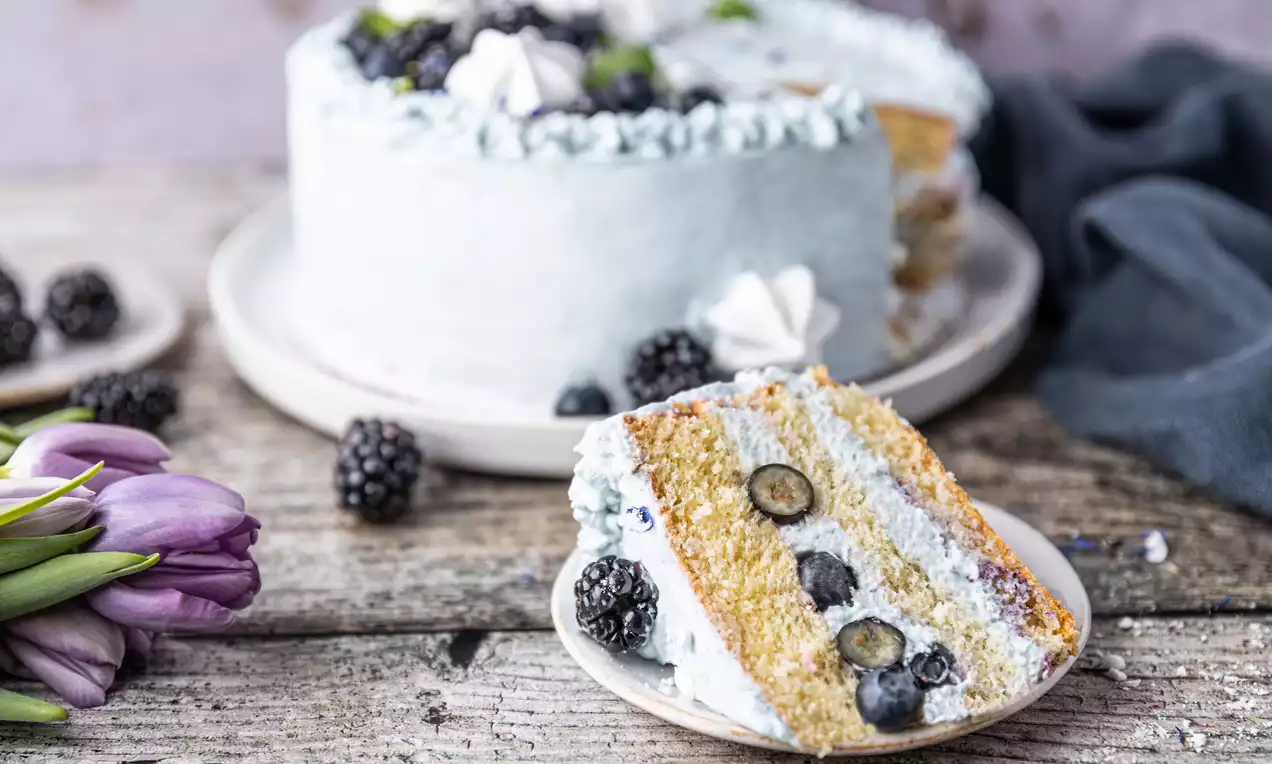 Niebieski tort warstwowy o kremowym wykończeniu, dekorowany jeżynami, borówkami, bezami i listkami mięty, na jasnym ceramicznym talerzu