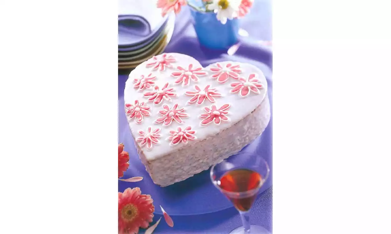 Gâteau en forme de cœur recouvert de glaçage blanc décoré de fleurs roses délicates sur une table violette avec fleurs et verre de liqueur