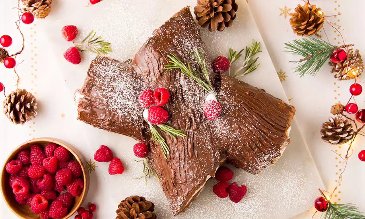 Tronco de Navidad con textura de corteza cubierta de crema marrón, decorado con frambuesas, ramitas verdes y azúcar glas sobre una bandeja
