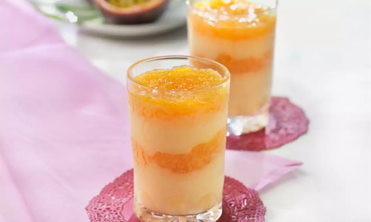 Capas cremosas de coco blanco y naranja brillante en vaso transparente sobre blonda rosa, con maracuyá partida al fondo sobre plato blanco