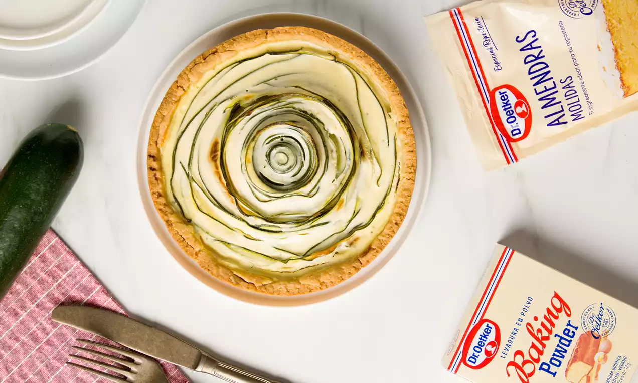 Tarta de masa dorada con finas láminas de calabacín verde dispuestas en espiral sobre un relleno claro en un plato blanco