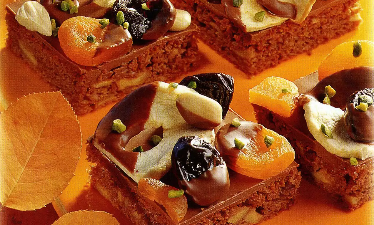 Carrés de gâteau aux noix brun doré garnis de fruits secs, pistaches et chocolat, présentés sur un fond orange avec des feuilles