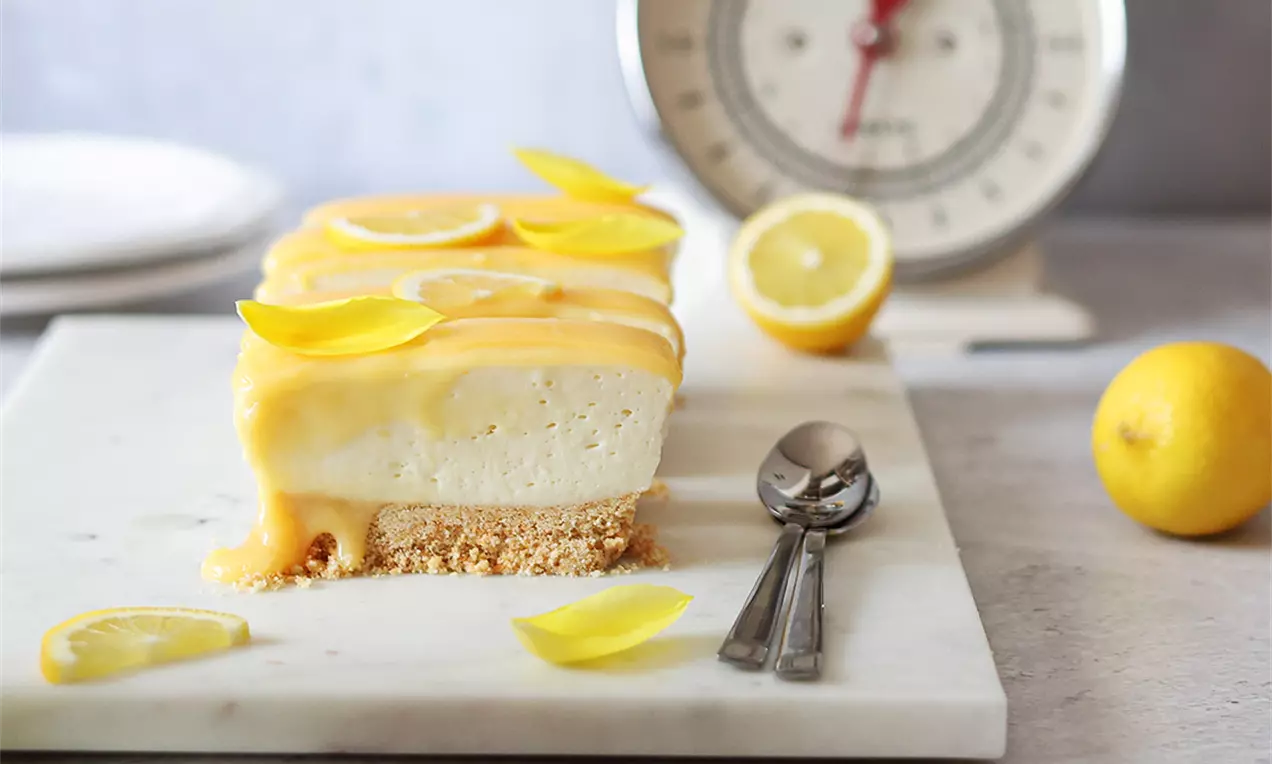 Правоъгълен лимонов сладкиш с кремообразна текстура върху хрупкава основа от трохи, покрит с жълт лимонов сос и свежи резени лимон