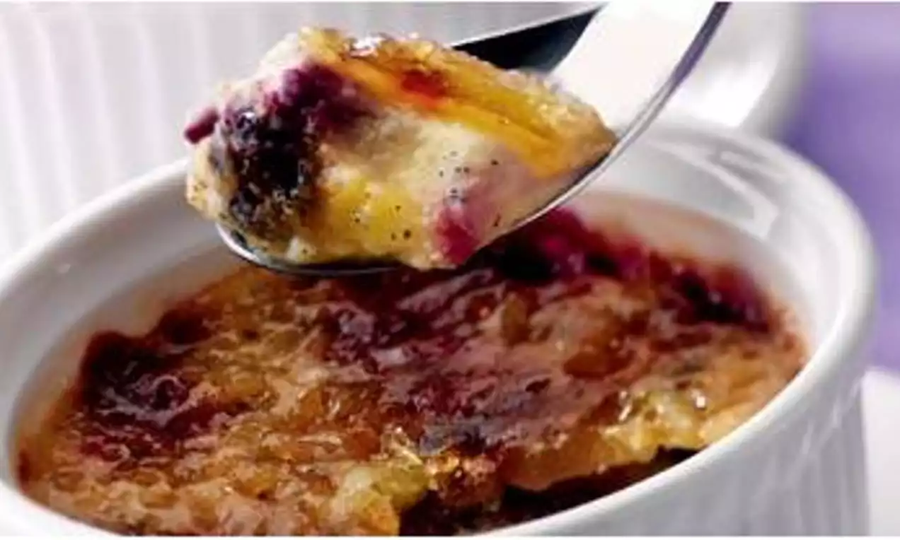 Blueberry crème brûlée in a white ramekin with golden caramelised topping, creamy interior dotted with purple berries, spoon lifting a portion