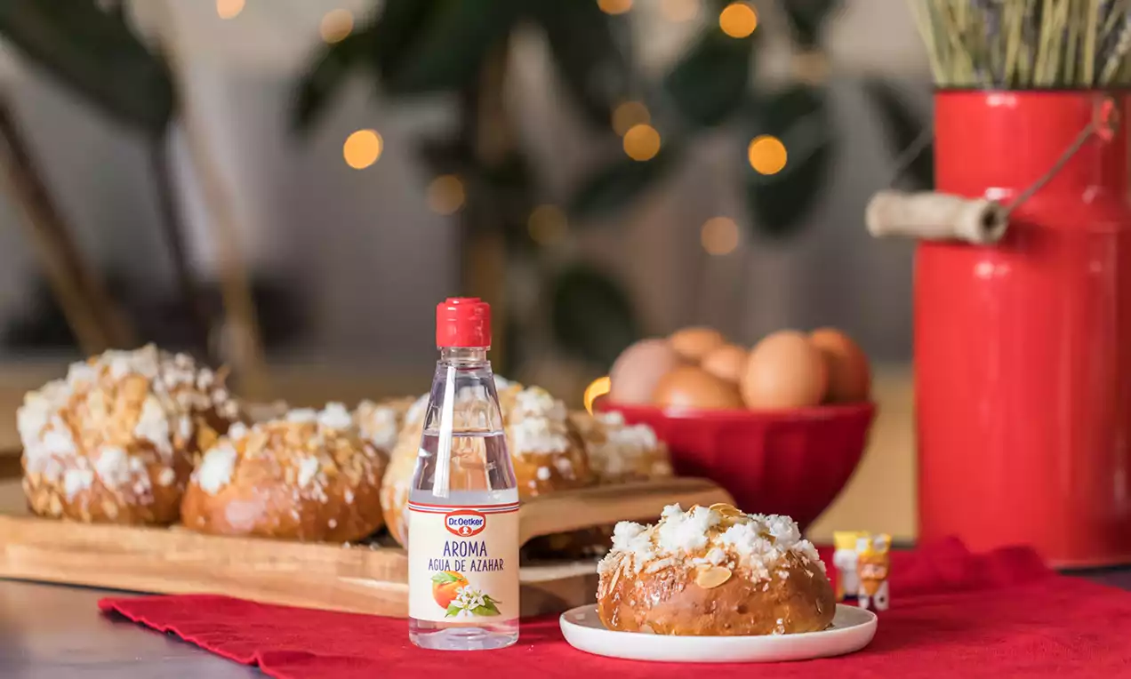 Mini roscón dorado y redondeado cubierto de azúcar granulada blanca sobre plato blanco, rodeado de ingredientes y decoración festiva