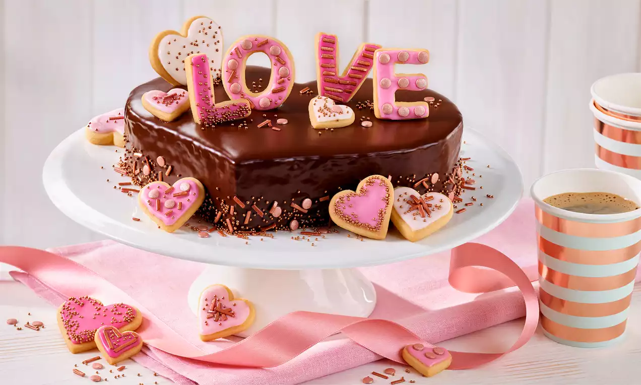 Herztorte mit glänzendem dunklen Schokoguss auf weißer Kuchenplatte, dekoriert mit rosa Keksen, Streuseln und einem love-Schriftzug