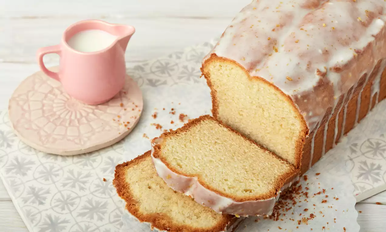 Zitronenkuchen mit heller Glasur und goldbraunem Teig, angeschnitten auf gemustertem Papier, daneben rosa Milchkännchen auf hellem Untergrund