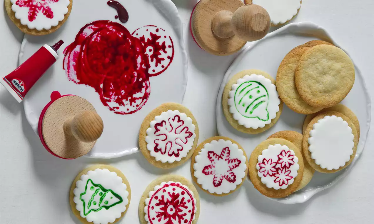 Mandlová kolečka světle zlatá s hladkou vrstvou bílého cukrového polevu, zdobená červenými a zelenými vánočními motivy na bílém talíři