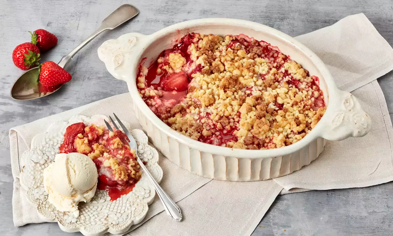 Erdbeer-Crumble mit goldbrauner Kruste und roter Fruchtfüllung in ovaler Backform, daneben Eis und Crumble auf Teller mit Löffel