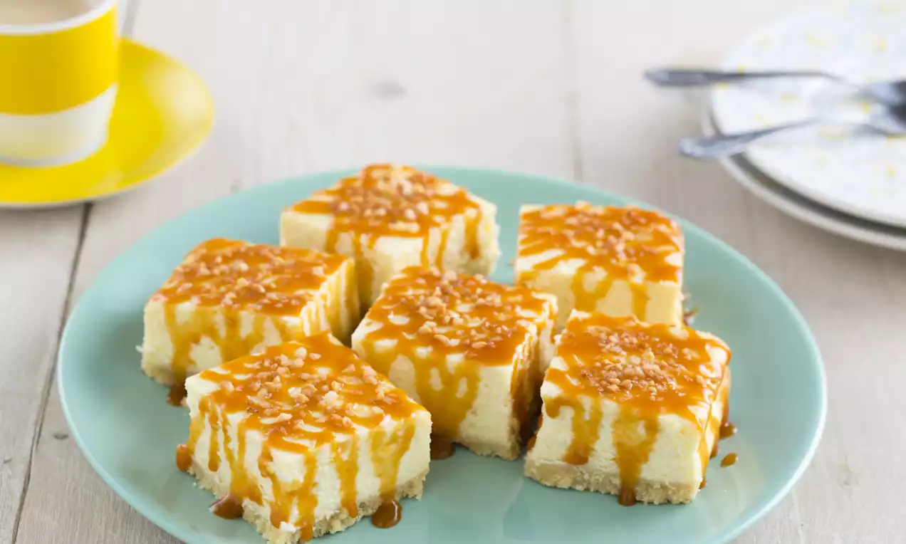 Blokjes lichtgele cheesecake met een kruimelbodem en glanzende stroop van karamel op een pastelblauw bord, met vorken en kopje op de achtergrond
