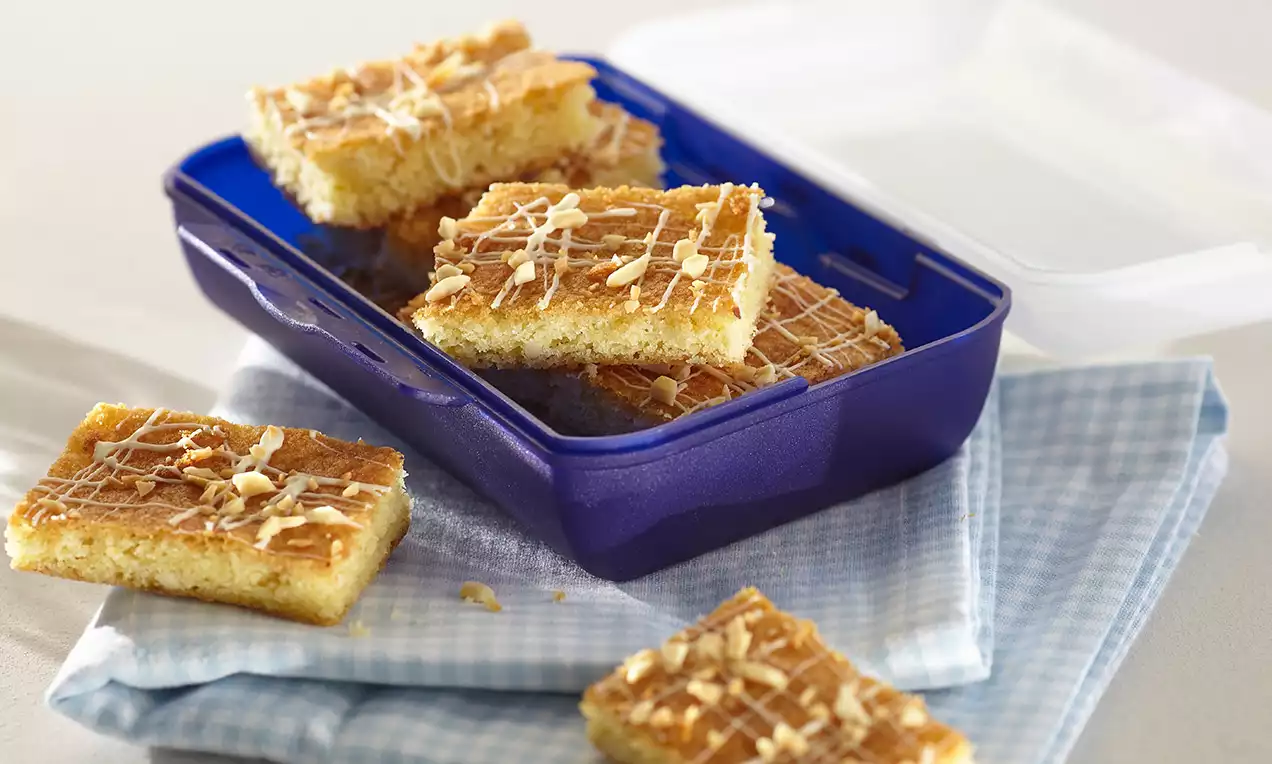 Blondies mit goldbrauner Kruste und heller Innenseite, rechteckig geschnitten, mit weißer Glasur und Mandelsplittern in einer blauen Dose auf kariertem Stoff